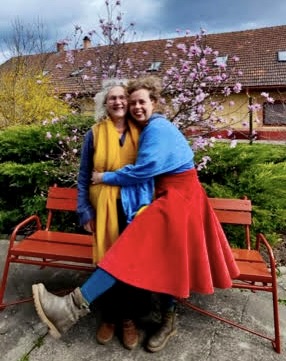 Two women smile and embrace each other outside in front of a blossoming tree.